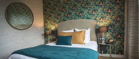 A neatly made bed with a teal blanket, a round mirror on the wall, and a floral wallpaper pattern.