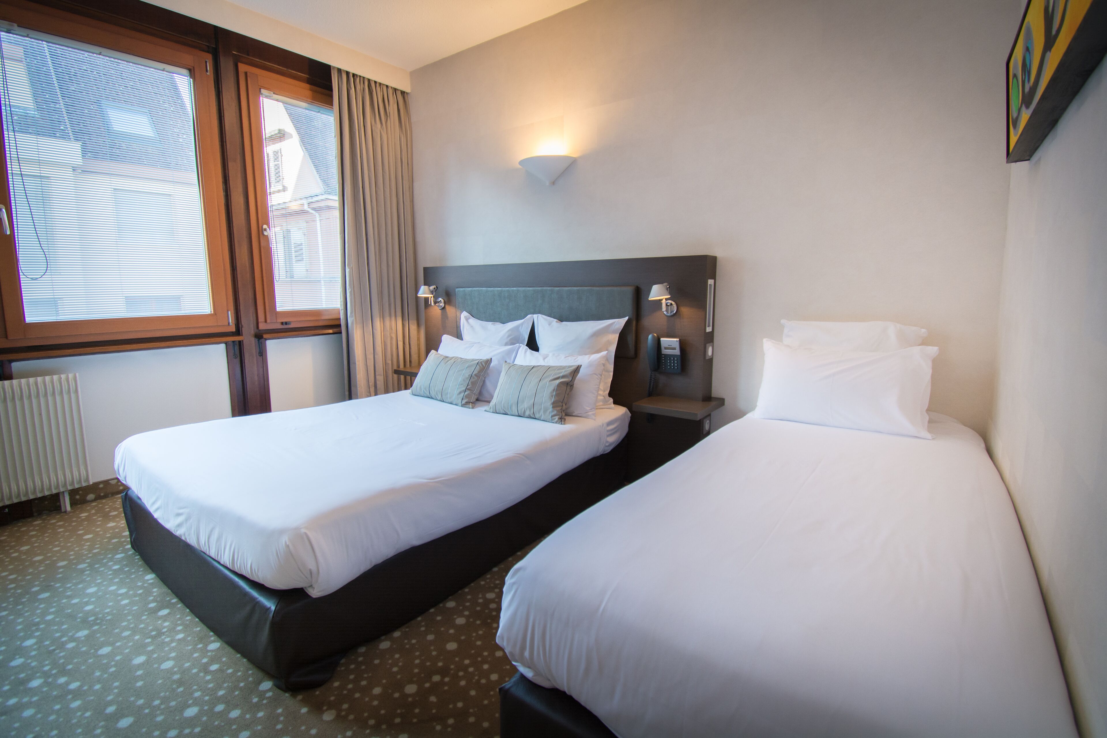 Quarto de hotel com duas camas de solteiro, uma janela grande com cortinas, luminária fixada na parede e radiador.
