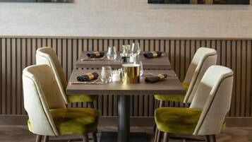 A dining area with a table set for two, chairs with green upholstery, and a wall-mounted light fixture.