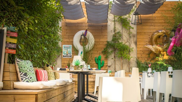 Un patio al aire libre con mesas de madera y sillas blancas, rodeado de follaje y un detalle decorativo de cactus.
