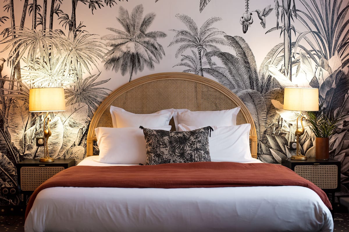 Een bed met een rond hoofdeinde, twee bedlampjes en een muurschildering met een tropisch thema.