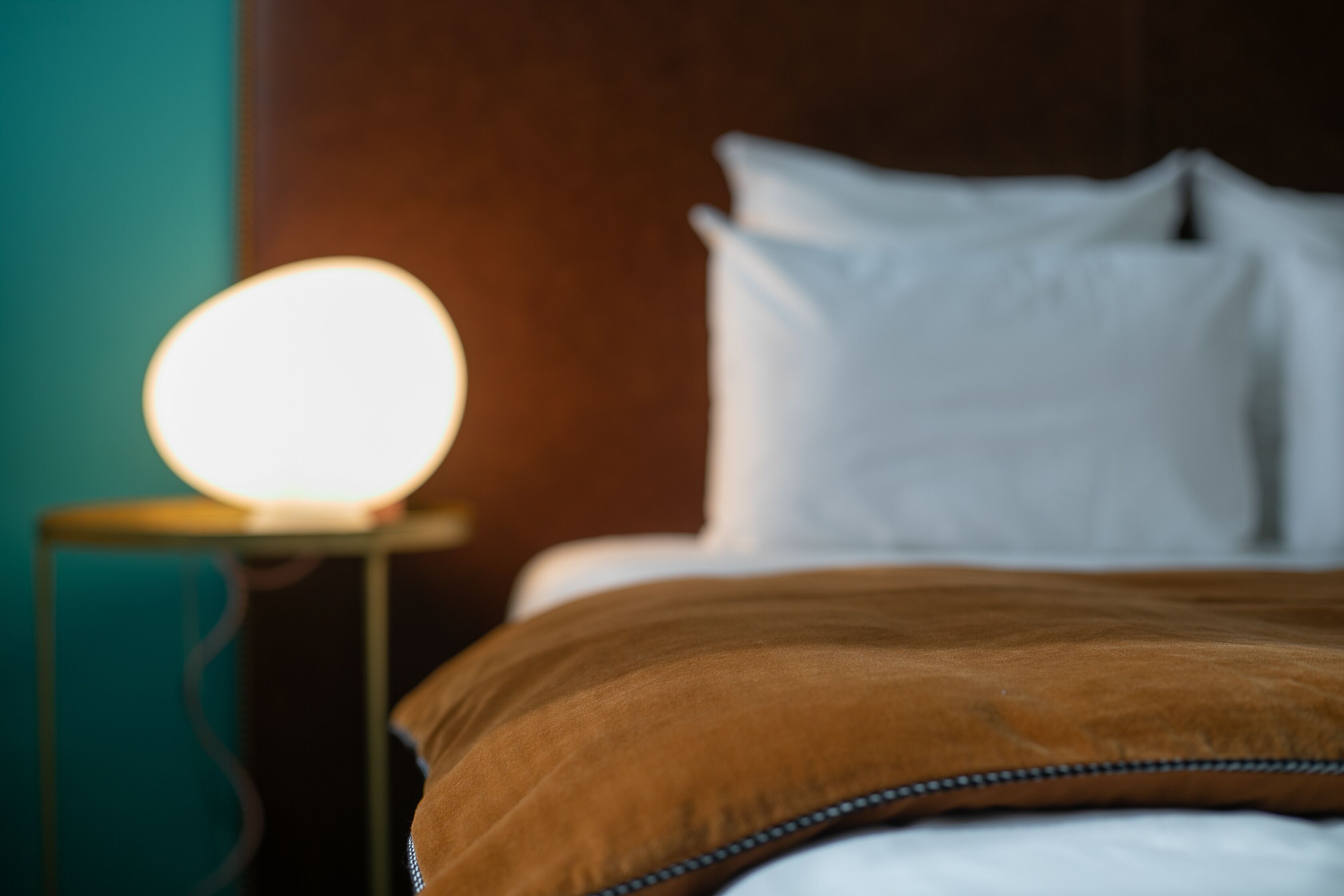 Un lit avec des oreillers blancs et une couverture marron, une lampe de chevet avec une ampoule qui émet une lumière douce, et un mur de couleur turquoise.