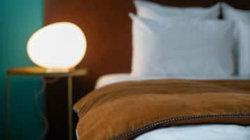 Una cama con almohadas blancas y una manta marrón, una lámpara de noche con una bombilla encendida, y una pared color turquesa.