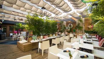 An outdoor dining area with wooden tables and chairs, surrounded by greenery and covered by large, beige canopies.