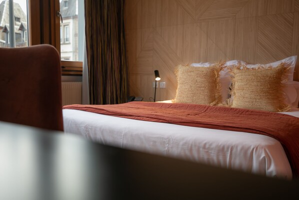 Habitación de hotel con cama, mesita de noche, lámpara y vista de edificios al exterior.