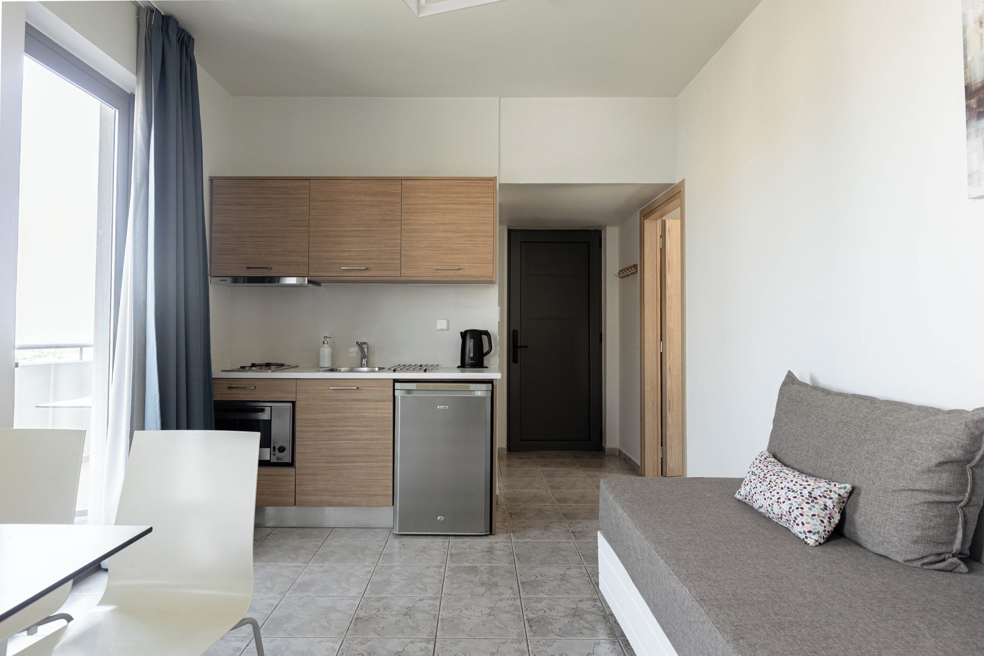 A compact kitchen with a dishwasher, a coffee maker, and a stove. There is a sofa with a patterned pillow and a window with curtains.