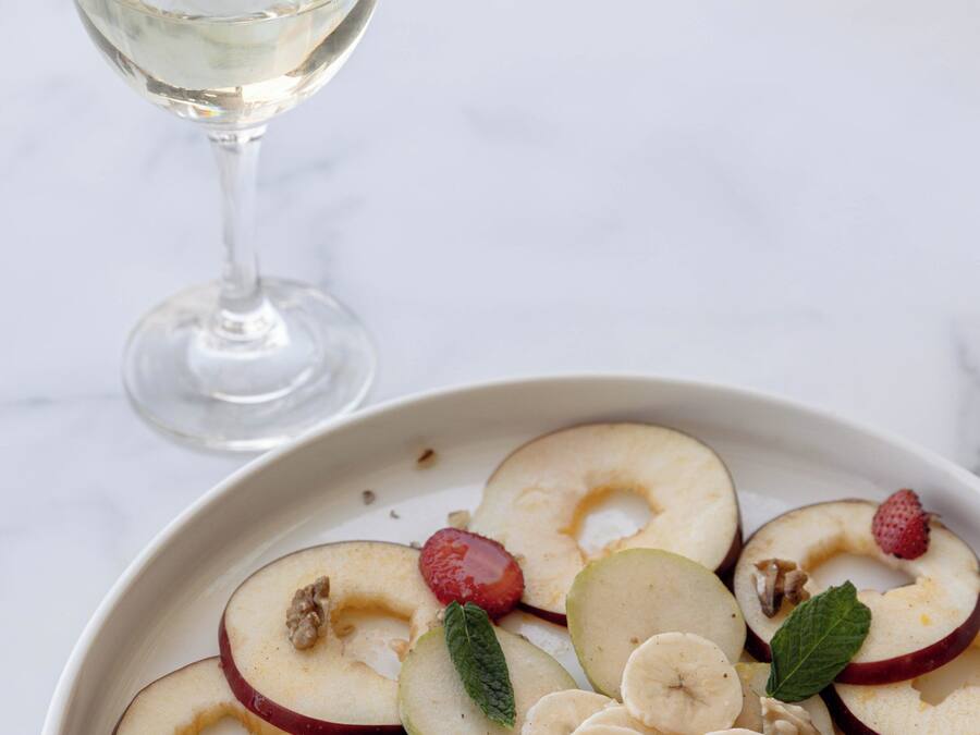 A plate of sliced apples, bananas, and strawberries with nuts and mint leaves, accompanied by a glass of white wine.