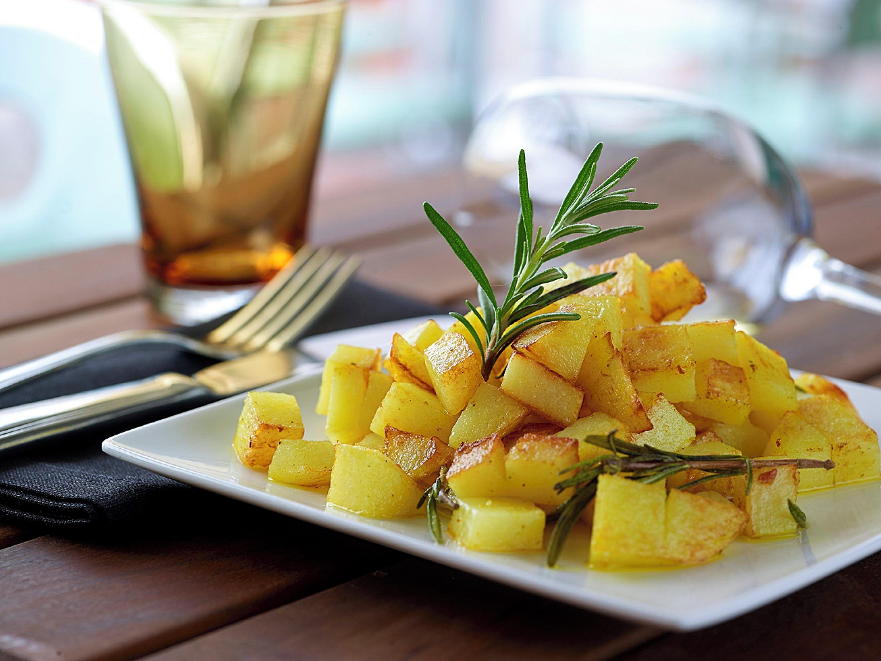 A plate of roasted potatoes garnished with rosemary, a fork, and a glass of beverage on a wooden table.