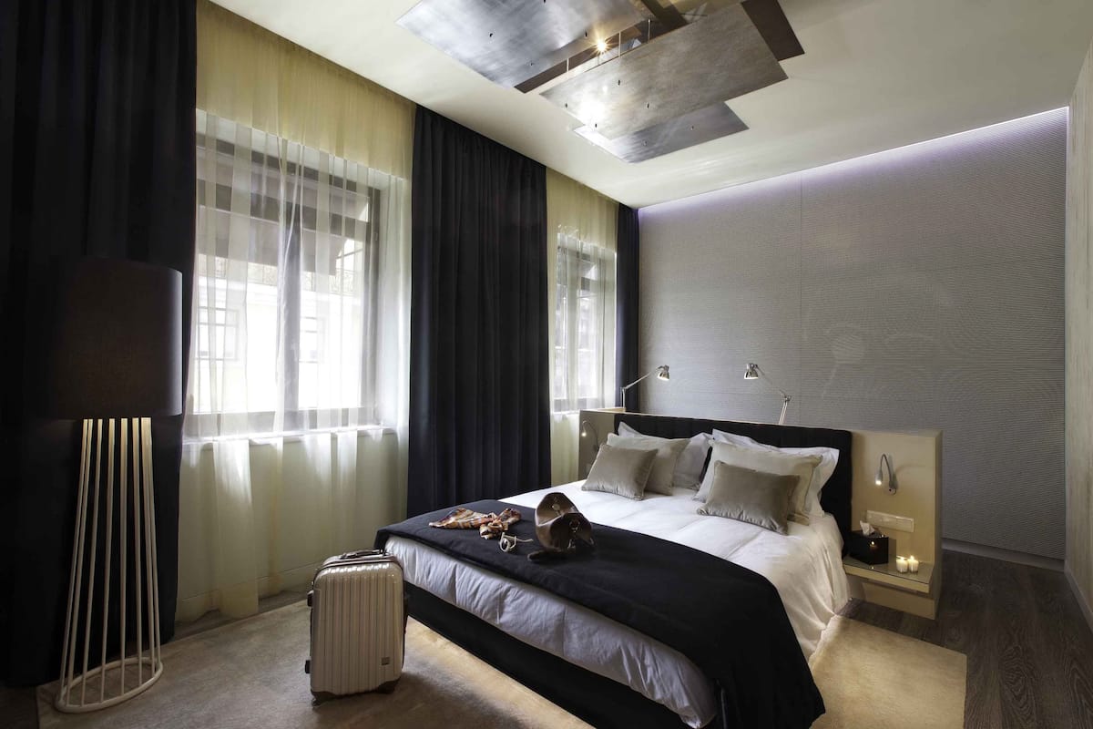 Una habitación de hotel moderna con una cama grande, mesitas de noche, una lámpara de pie y una maleta en el suelo.