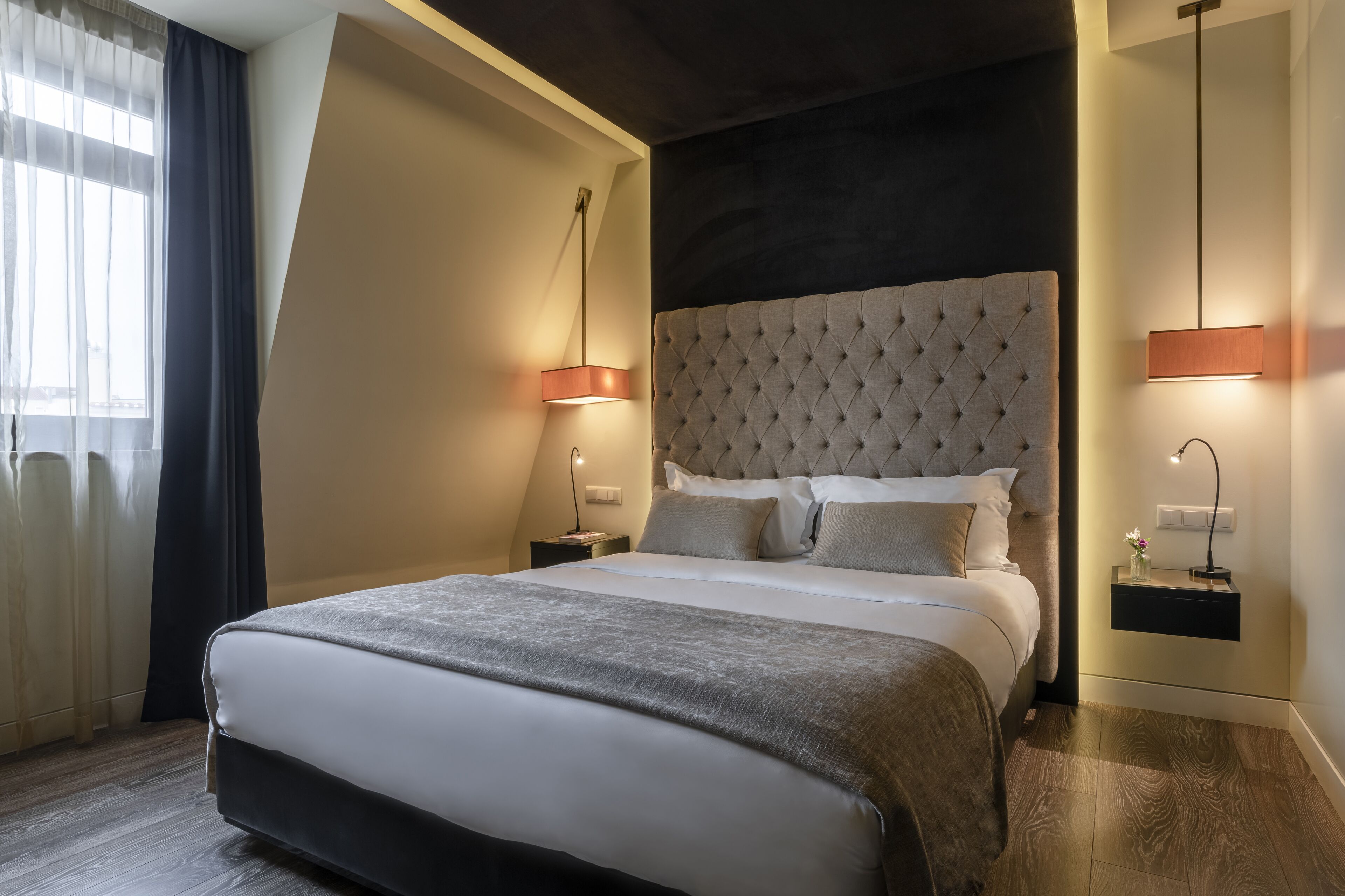 Habitación de hotel moderna con una cama grande, cabecera capitonada, mesitas de noche con lámparas y vistas a la ciudad a través de cortinas translúcidas.