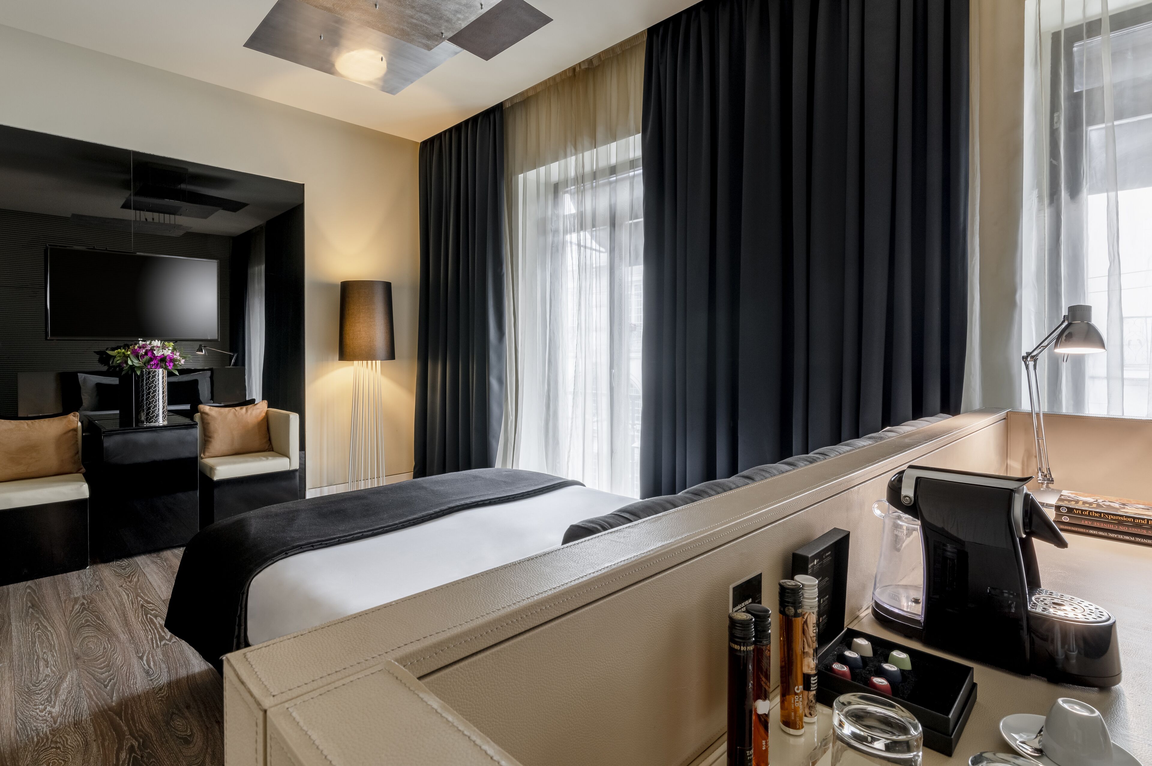 A modern hotel room with a large bed, a desk with a coffee maker, a TV, and a large window with curtains.