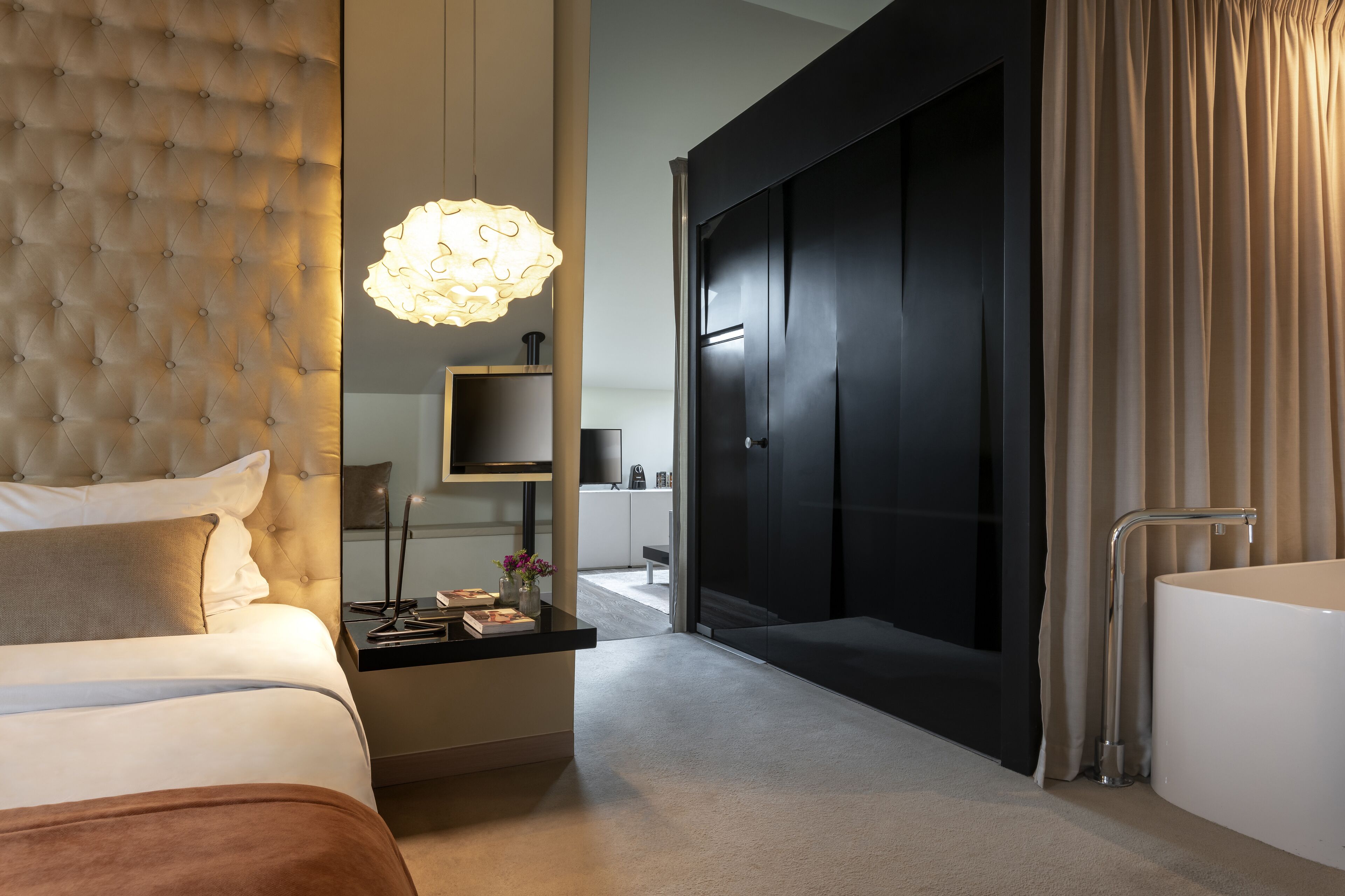 Una habitación de hotel moderna con cabecera capitonada, una mesita de noche con lámpara, un espejo grande y una puerta corredera que da a un baño.