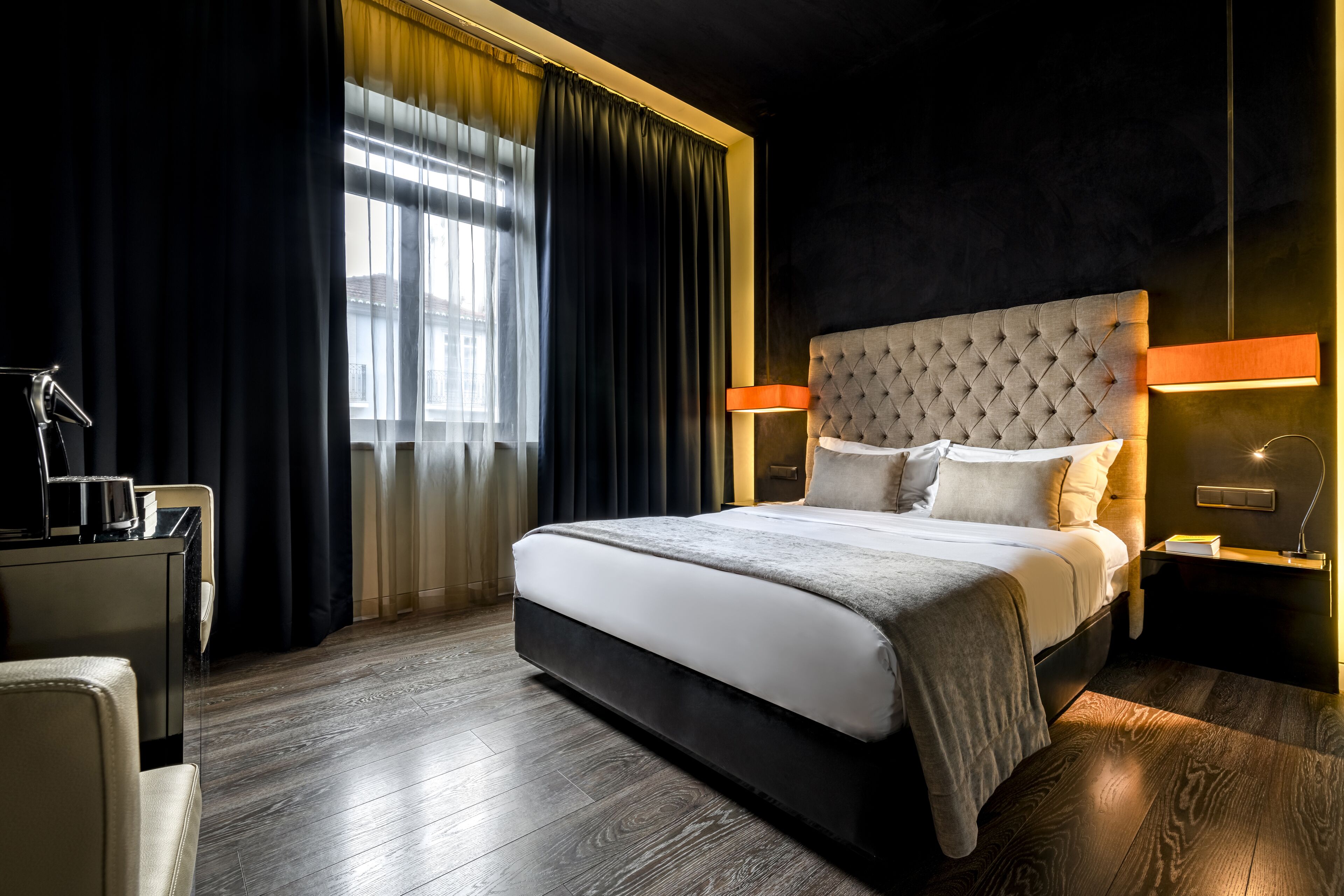 A modern hotel room with a large bed, dark curtains, and a nightstand with a lamp.