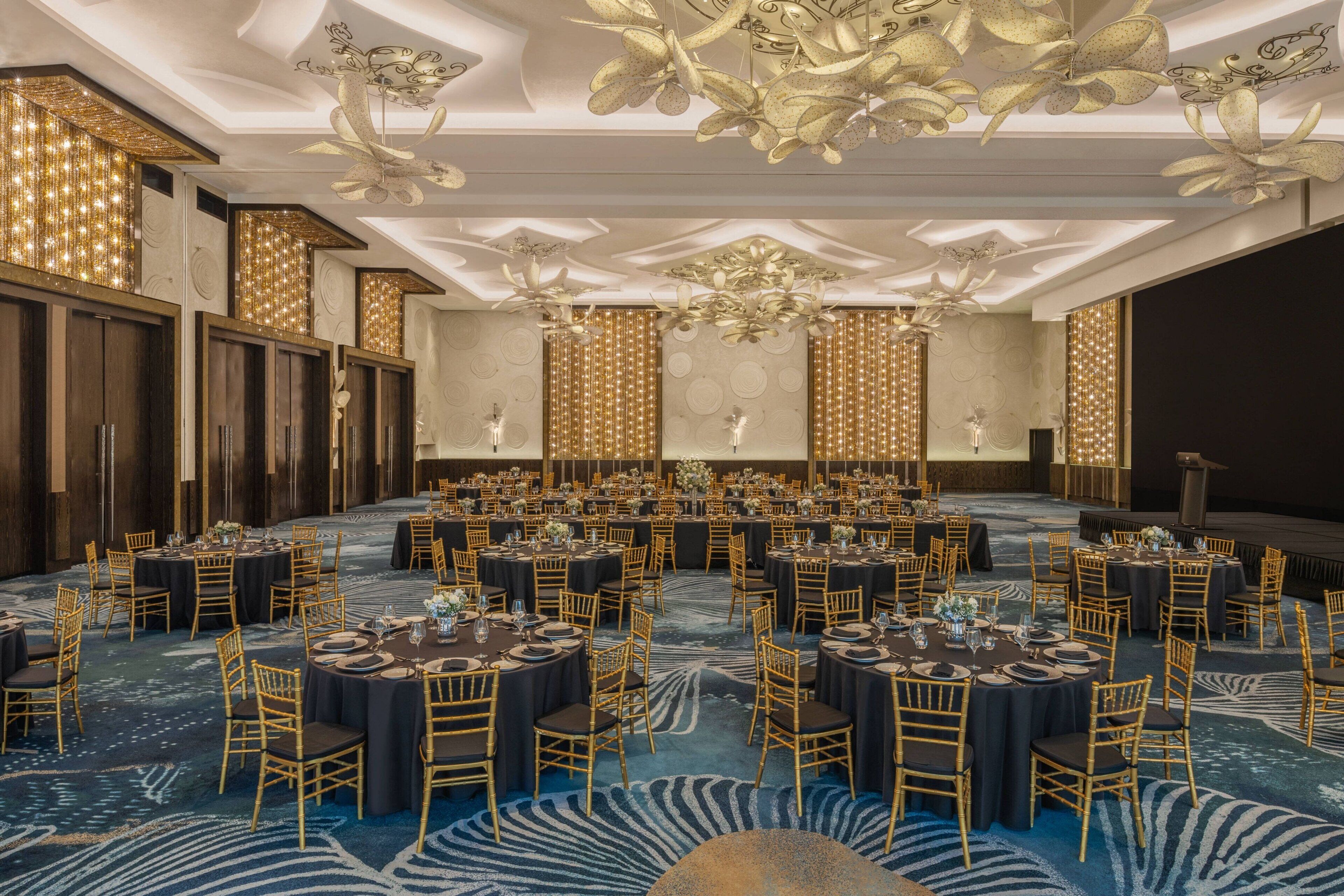 A large banquet hall with round tables, gold chairs, and a blue patterned carpet.
