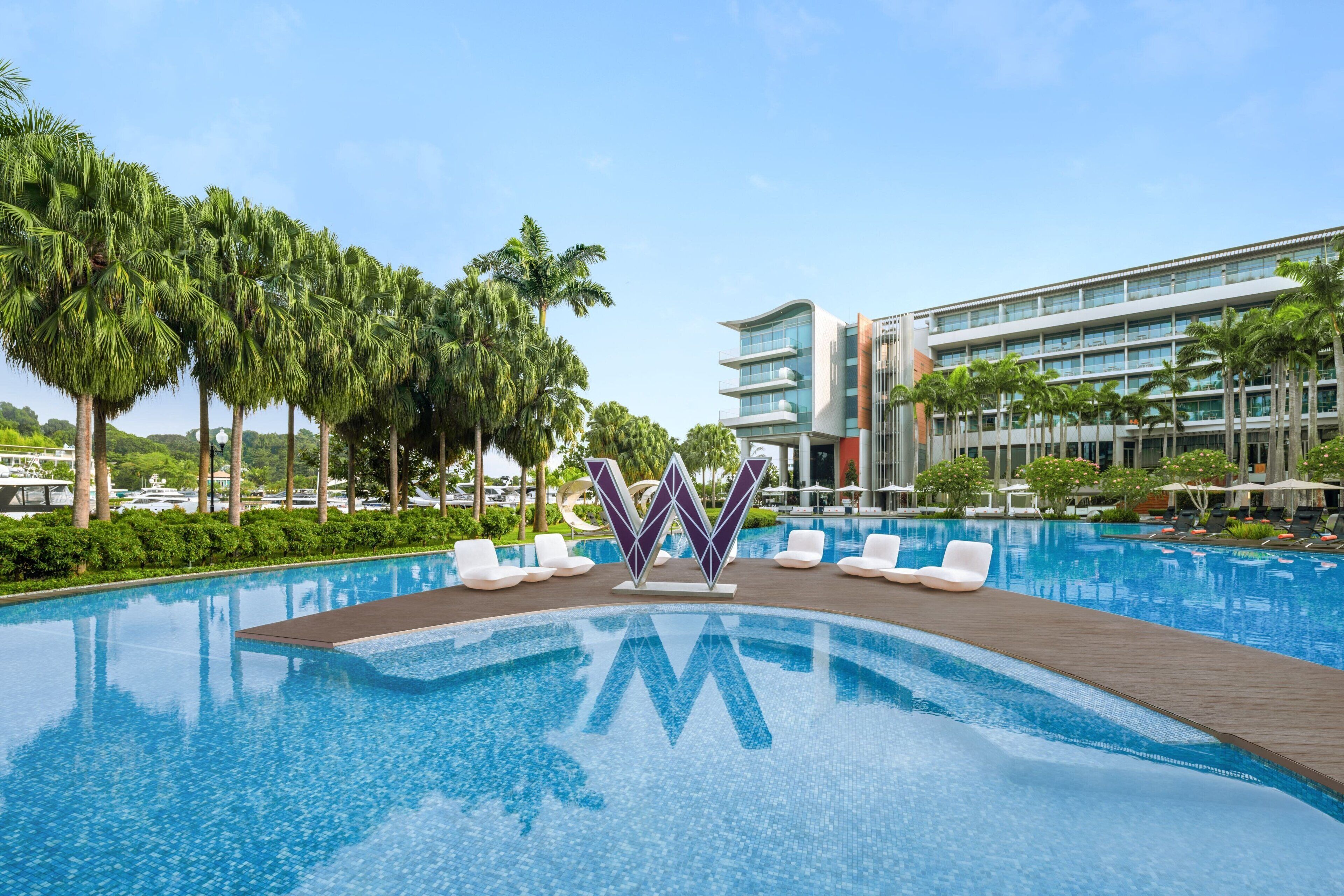 A swimming pool with lounge chairs, palm trees, and a modern building in the background.