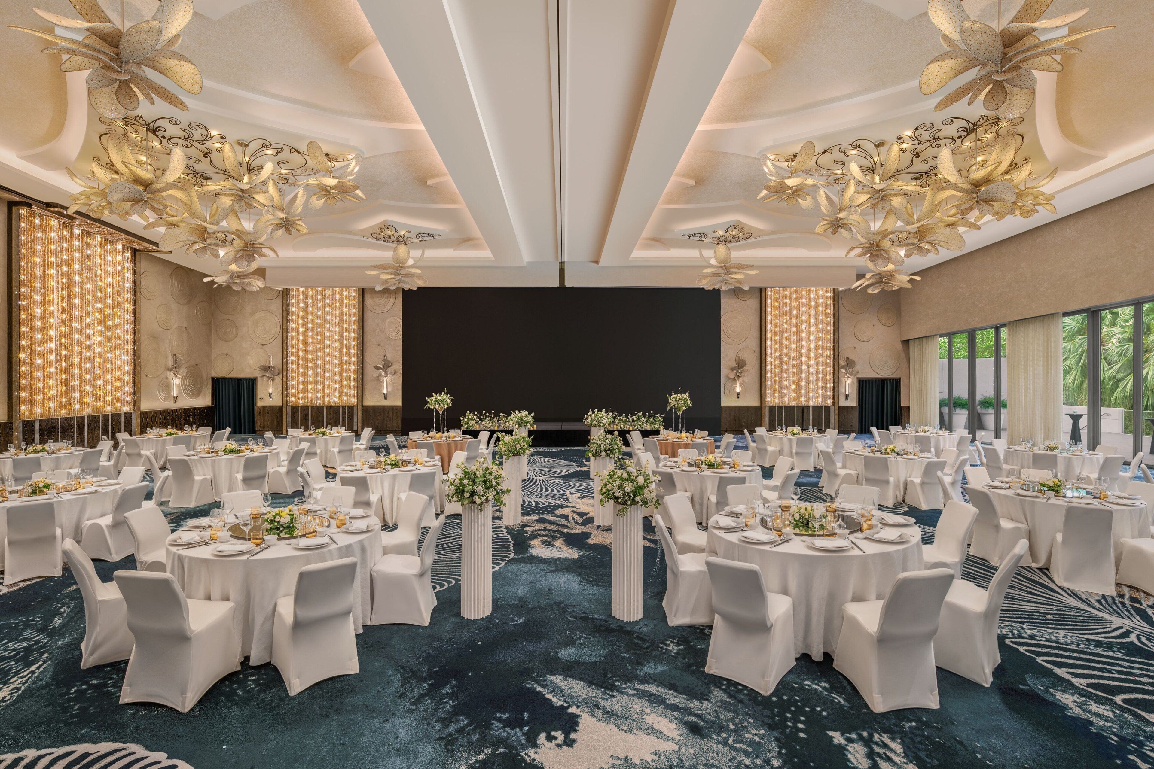 Un amplio salón de banquetes con mesas redondas, sillas blancas y centros de mesa florales.