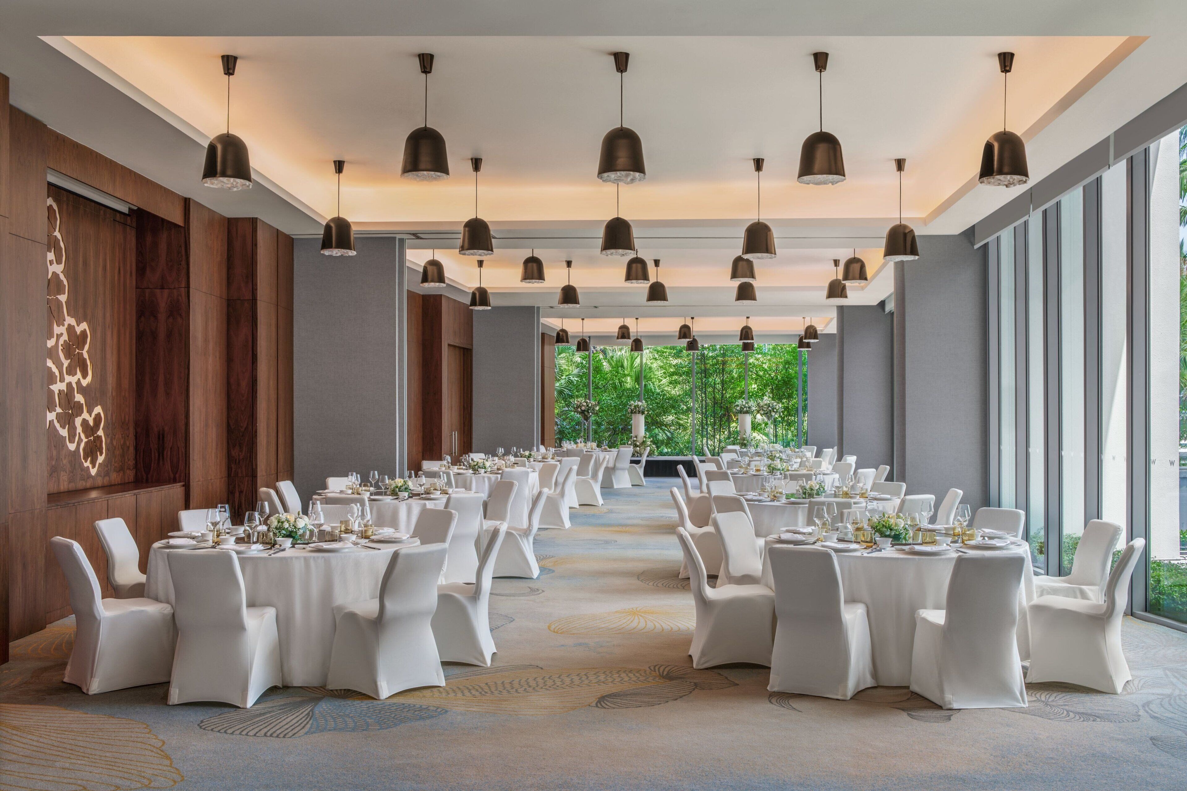 Un amplio salón de banquetes con varias mesas redondas dispuestas para comer, sillas blancas e iluminación colgante.