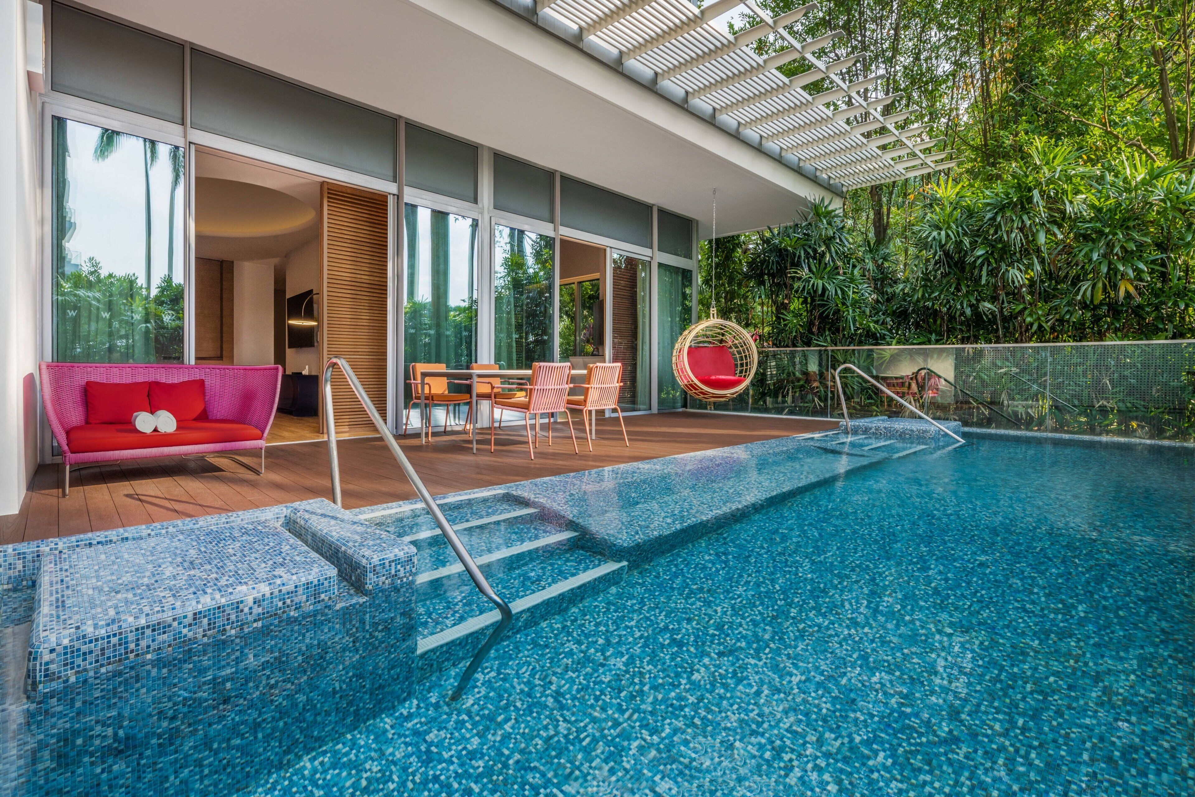Ein moderner Poolbereich im Innenbereich mit einer verglasten Terrasse, einem roten Sofa und einem Essplatz.
