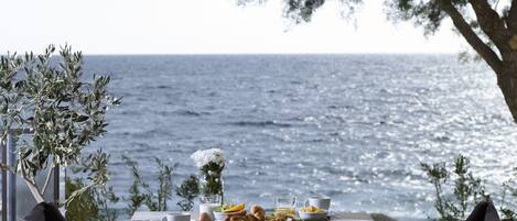 A table set for breakfast with a view of the sea, including plates, cups, and a vase of flowers.