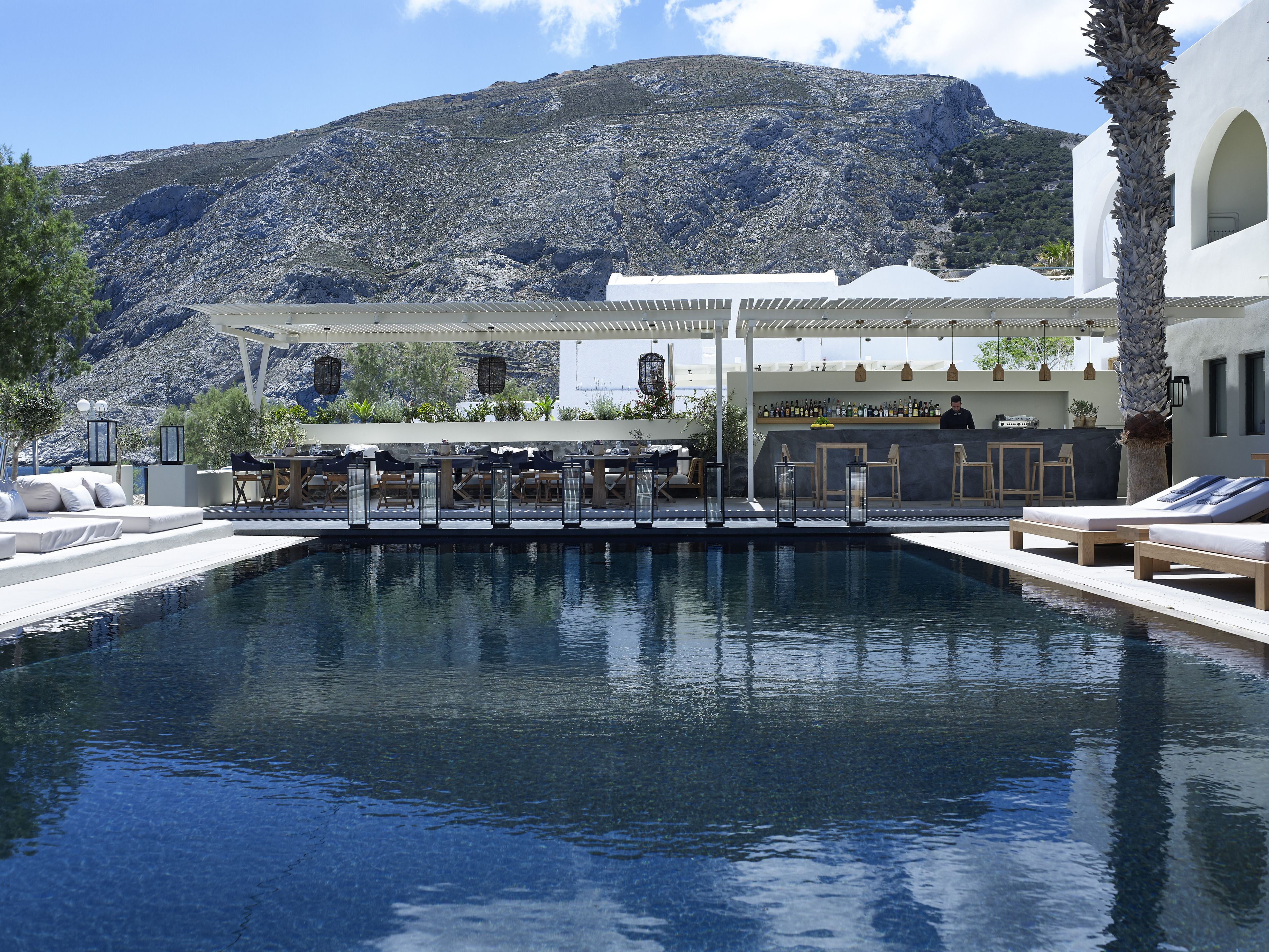 Zona de piscina con sillones de descanso y un bar, con montañas de fondo.