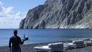 A beach with a rocky cliff, a man carrying a tray with bottles, and outdoor seating. - Bellonias Villas (Santorini)
