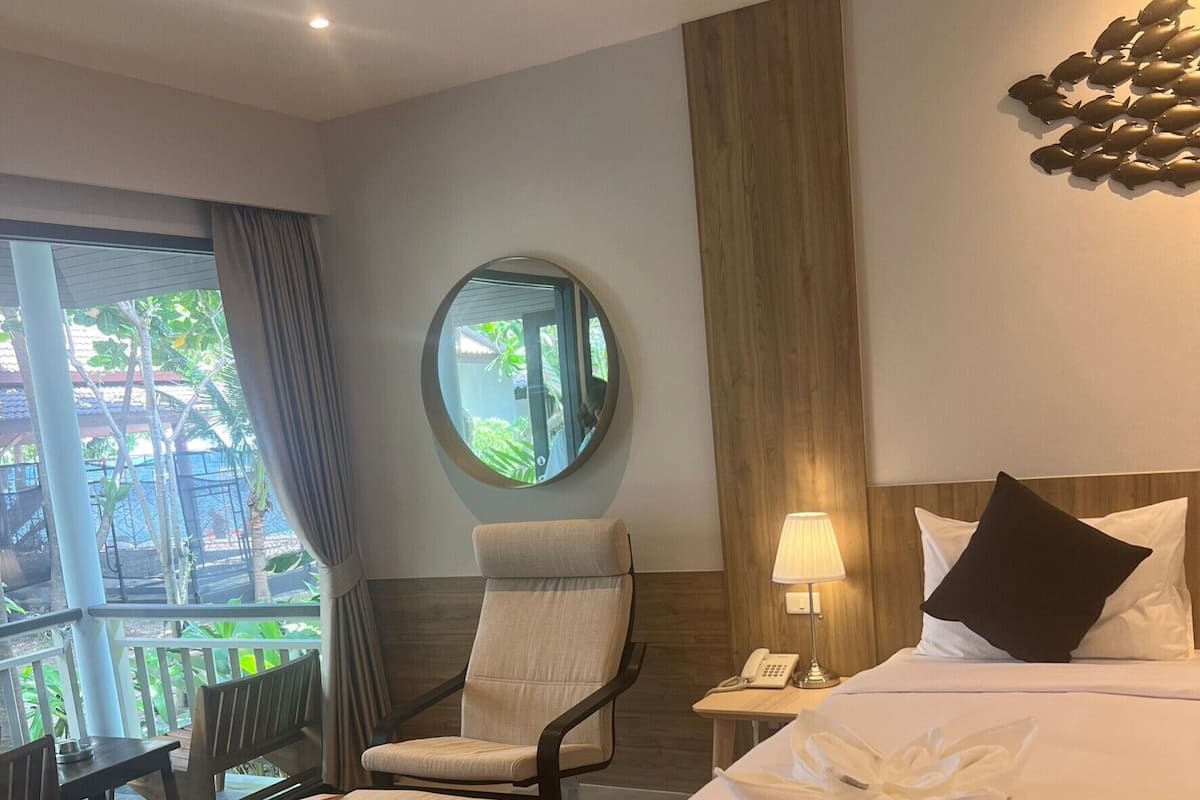 Habitación de hotel con cama, una silla, un espejo redondo, una mesita de noche y vistas al exterior.
