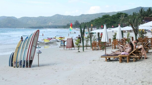 A beach with surfboards lined up, beach chairs, umbrellas, and a view of the sea and hills in the background. - Evergreen Resort (Koh Samui)