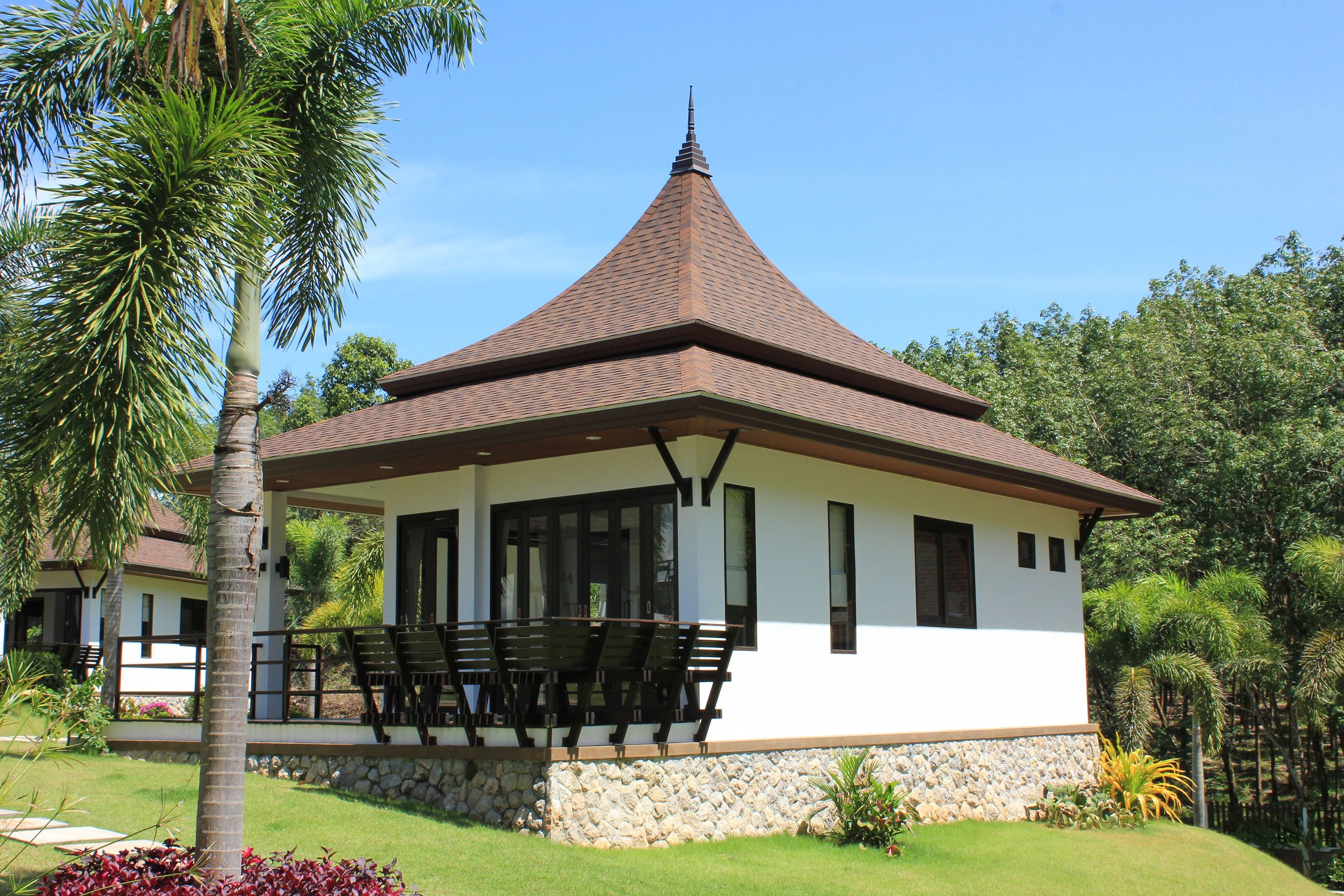 A white building with a brown roof and stone base, surrounded by greenery.