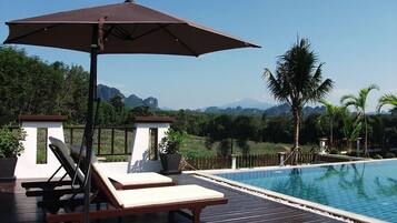 A poolside area with lounge chairs and an umbrella, surrounded by a wooden deck and a view of mountains and palm trees.