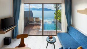 A modern living room with a large TV, a blue sofa, a wooden coffee table, and a small side table with fruit. A sliding glass door leads to a balcony with a pool and a view of the sea and mountains. - Kalima Resort & Spa, Phuket (Patong)