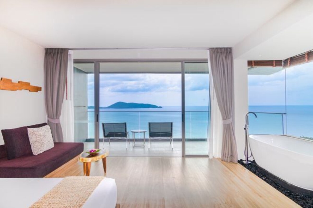Habitación de hotel con una cama grande, un sofá, una mesita, una bañera y un balcón con vistas al mar.