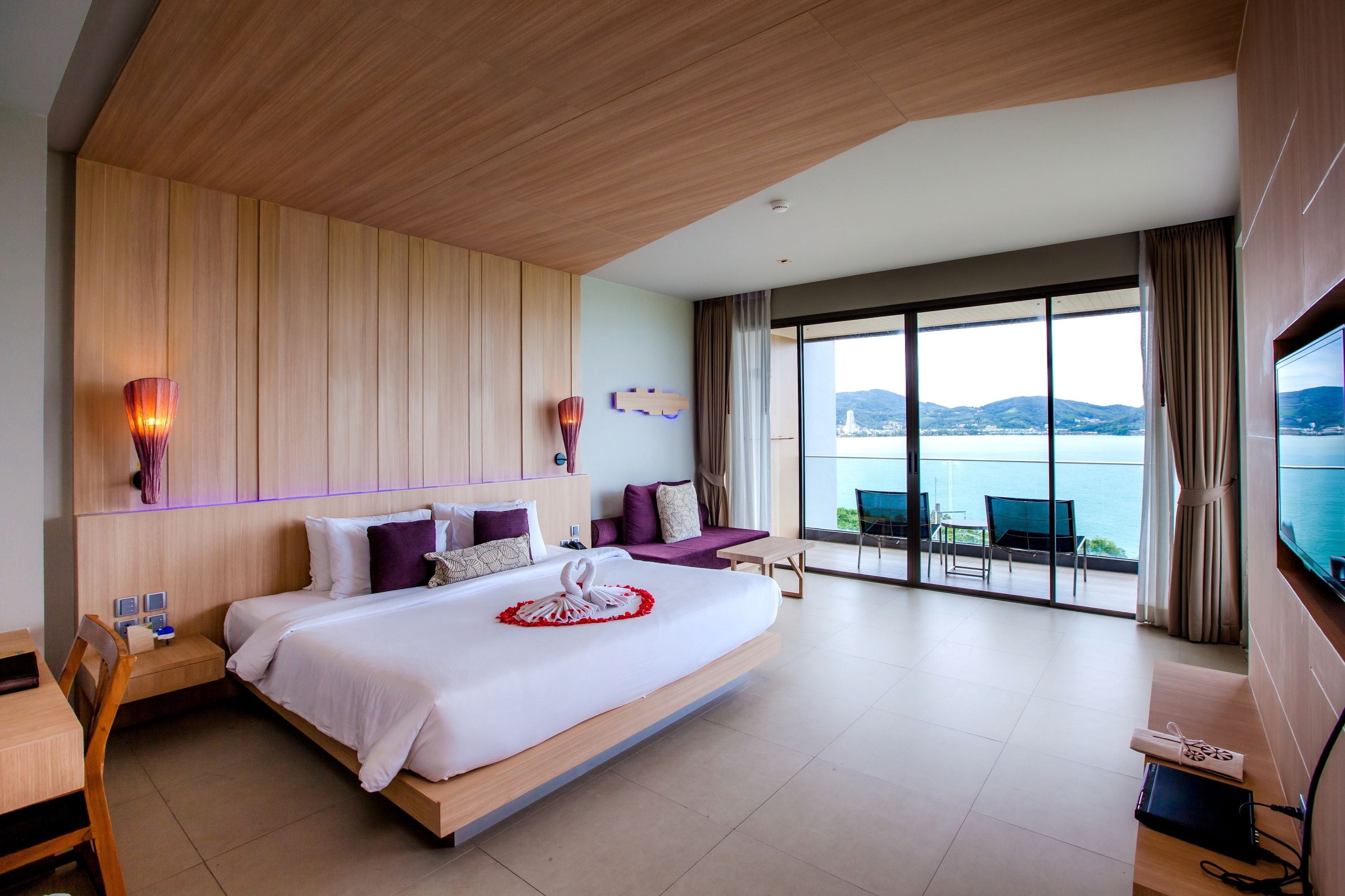 A modern hotel room with a large bed, a TV, a desk, and a balcony with a view of the sea.