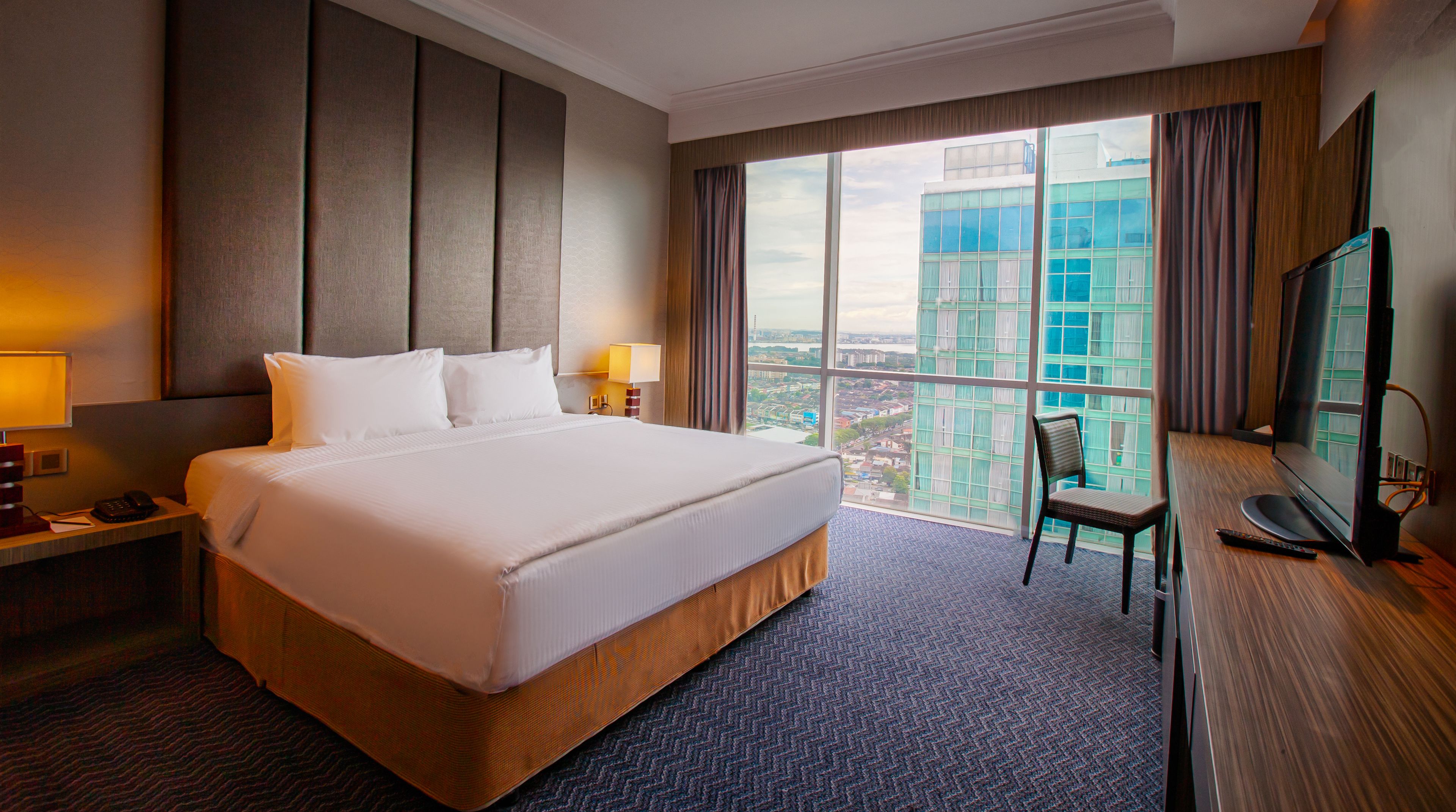 A hotel room with a large bed, a desk with a computer, a chair, and a view of a cityscape through large windows.