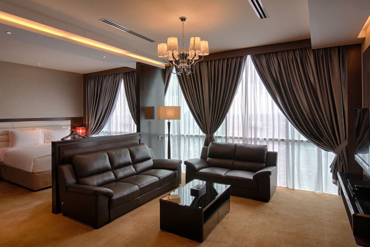 A modern hotel room with a king-sized bed, a dark leather sofa, a glass coffee table, and a large window with curtains.