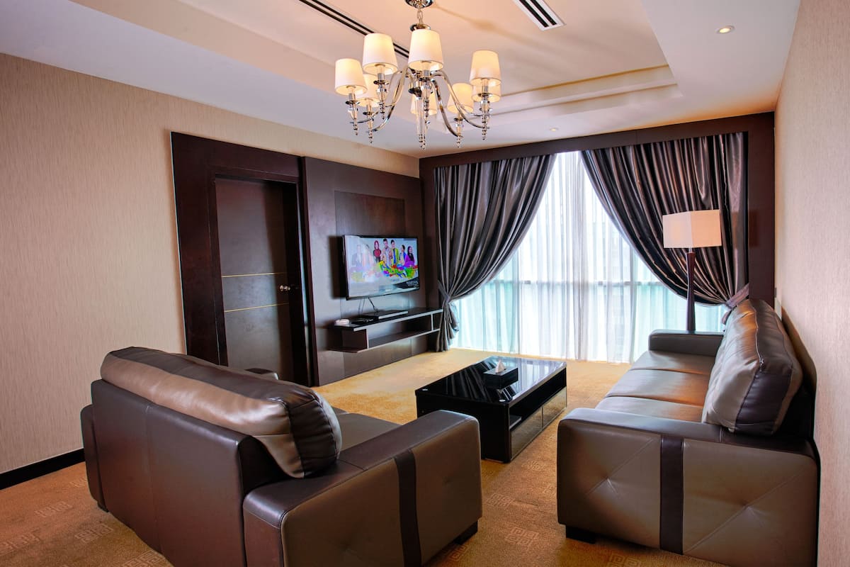 A modern hotel room with leather sofas, a flat-screen TV, a chandelier, and a large window with curtains.