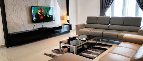Ein modernes Wohnzimmer mit einem großen Flachbildfernseher an einer Marmorwand, einem grauen Sofa, einem Glastisch und einer Lampe auf einem Beistelltisch.
