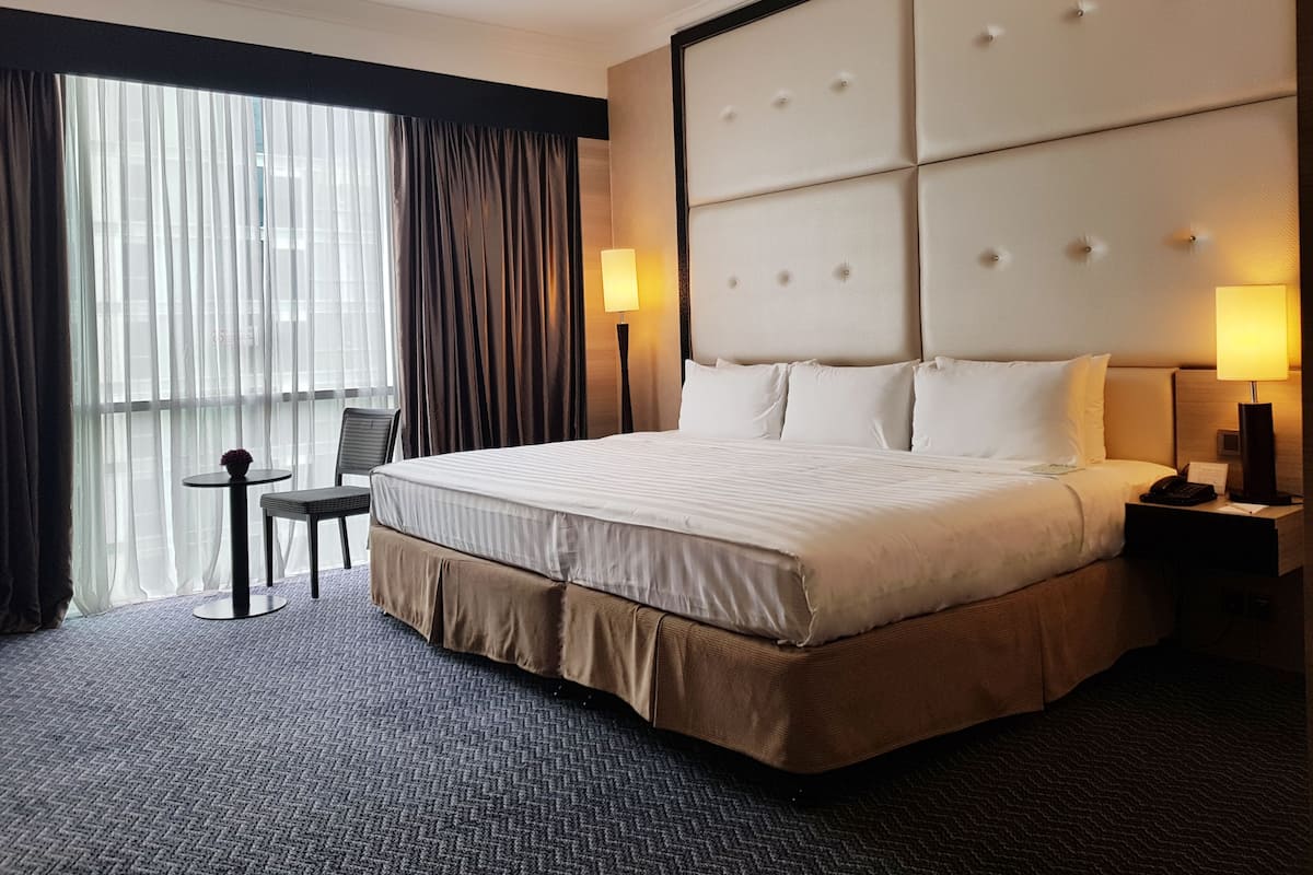 A hotel room with a large bed, a bedside table, a chair, a lamp, and a view of the city through the window.