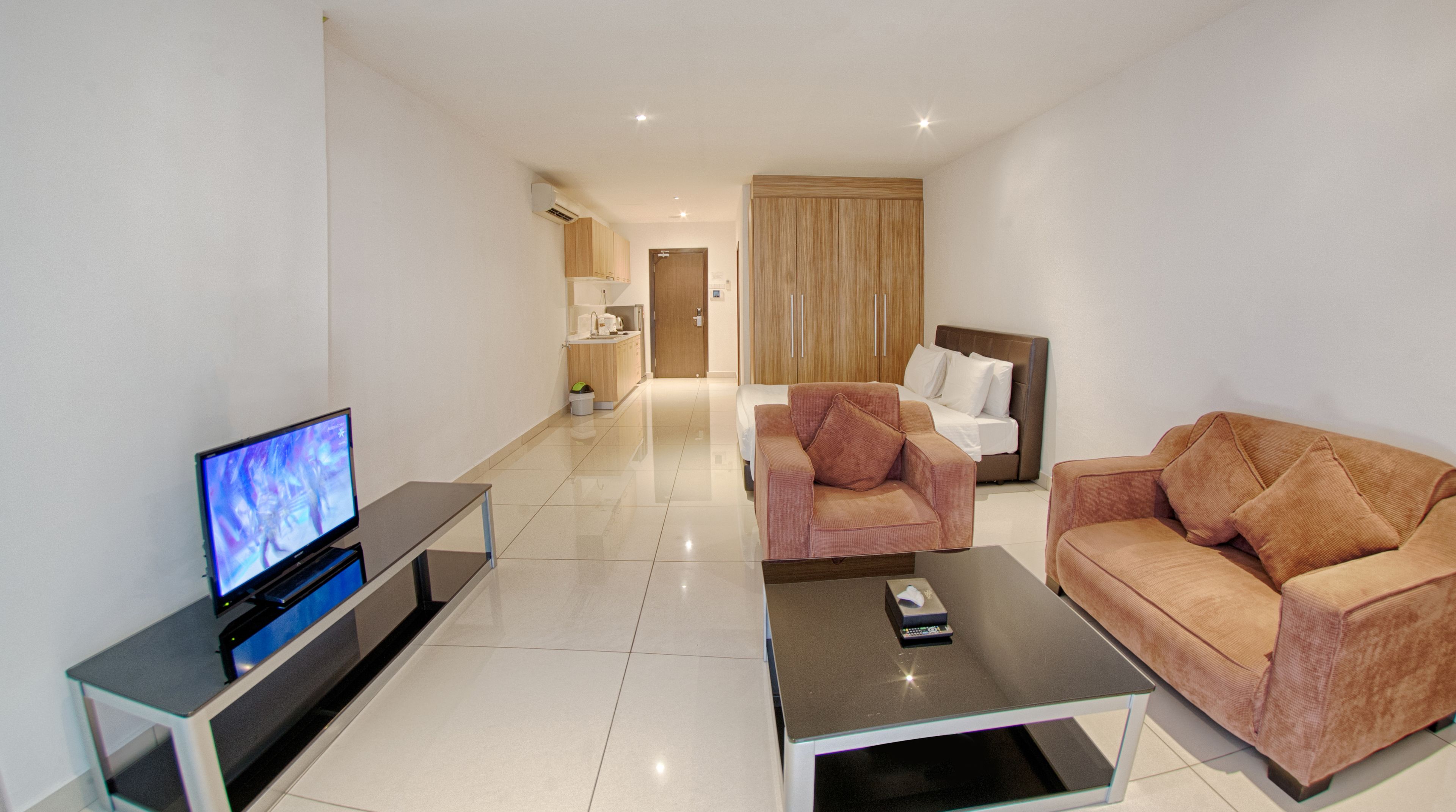 A modern living room with a flat-screen TV, a glass coffee table, two sofas, and a kitchen area in the background.
