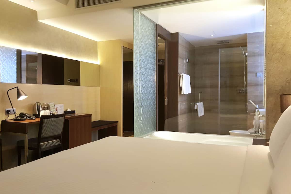 A modern hotel room with a large bed, a desk with a lamp, a shower area, and a bathroom with a bathtub.