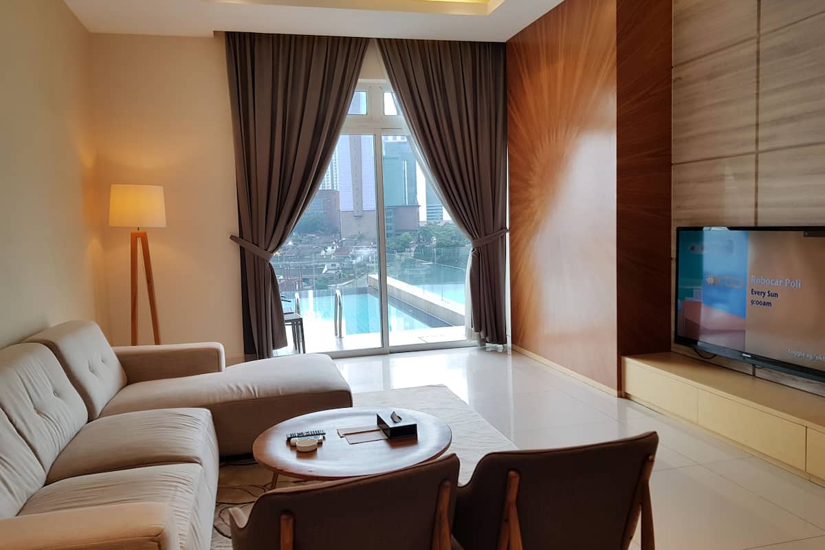 A modern hotel room with a sofa, chairs, a small table, a TV, and a view of a cityscape through large windows.