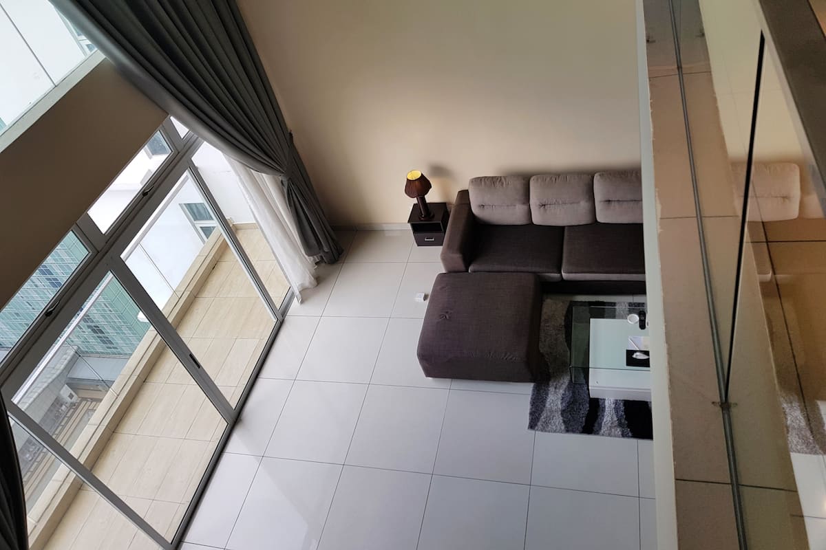 A modern living room with a sectional sofa, a small table, and a lamp. Large windows provide a view of the cityscape.