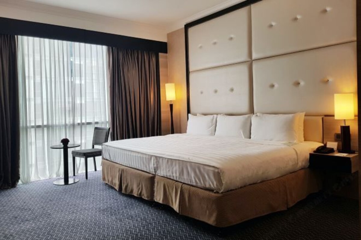 A hotel room with a large bed, two bedside tables, a small table and chair, and a large window with curtains.
