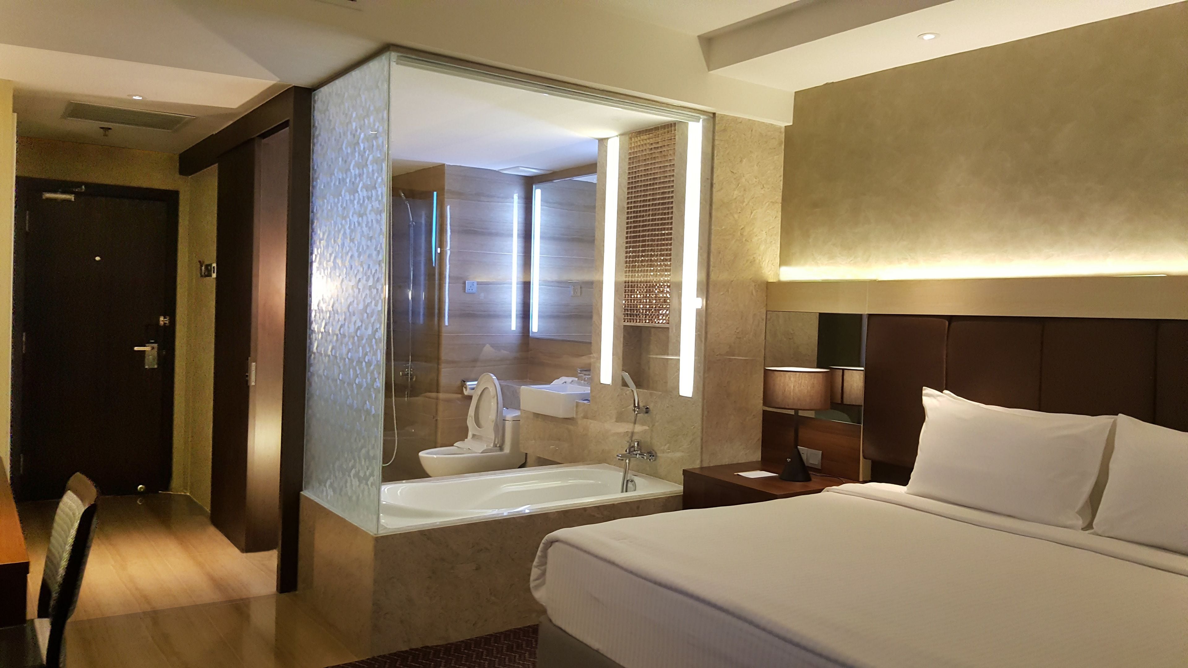 A hotel room with a large bed, a bathroom with a bathtub, a mirror, and a nightstand with a lamp.