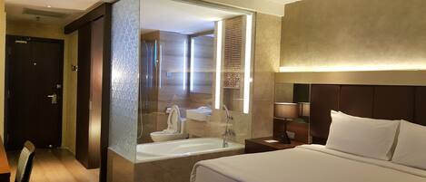 A hotel room with a large bed, a bathroom with a bathtub, a mirror, and a nightstand with a lamp.
