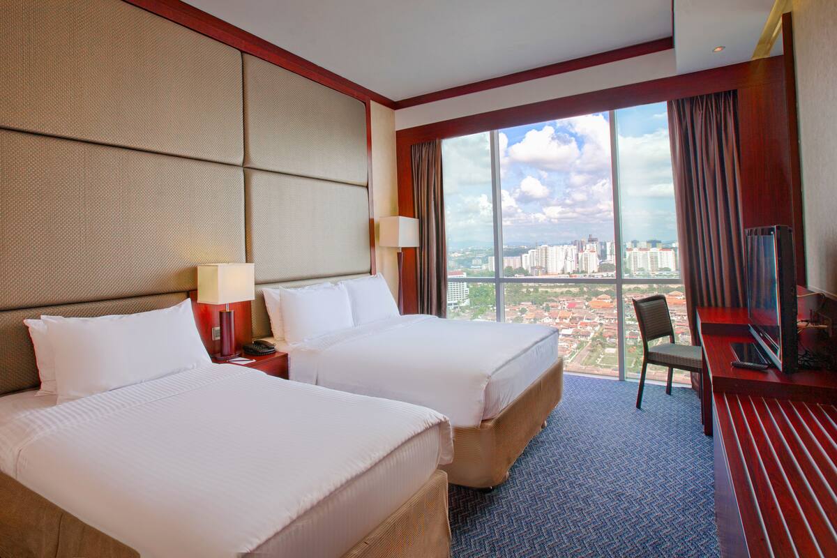 A hotel room with two beds, a desk with a TV, and a large window offering a city view.