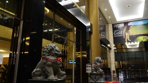 Un moderno vestíbulo de hotel con un mostrador de recepción, dos estatuas de leones y una pantalla grande mostrando un anuncio de simulador de golf para interior.