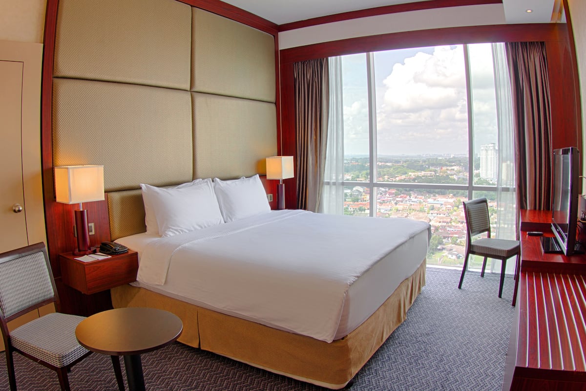 A hotel room with a large bed, two bedside tables with lamps, a chair, a desk with a TV, and a view of a cityscape through large windows.