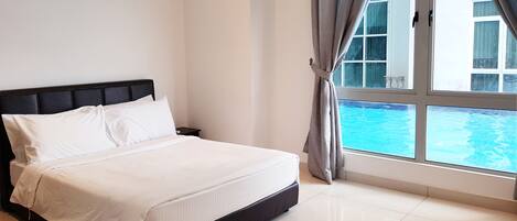 A neatly made bed with white linens in a hotel room with a large window overlooking a swimming pool.