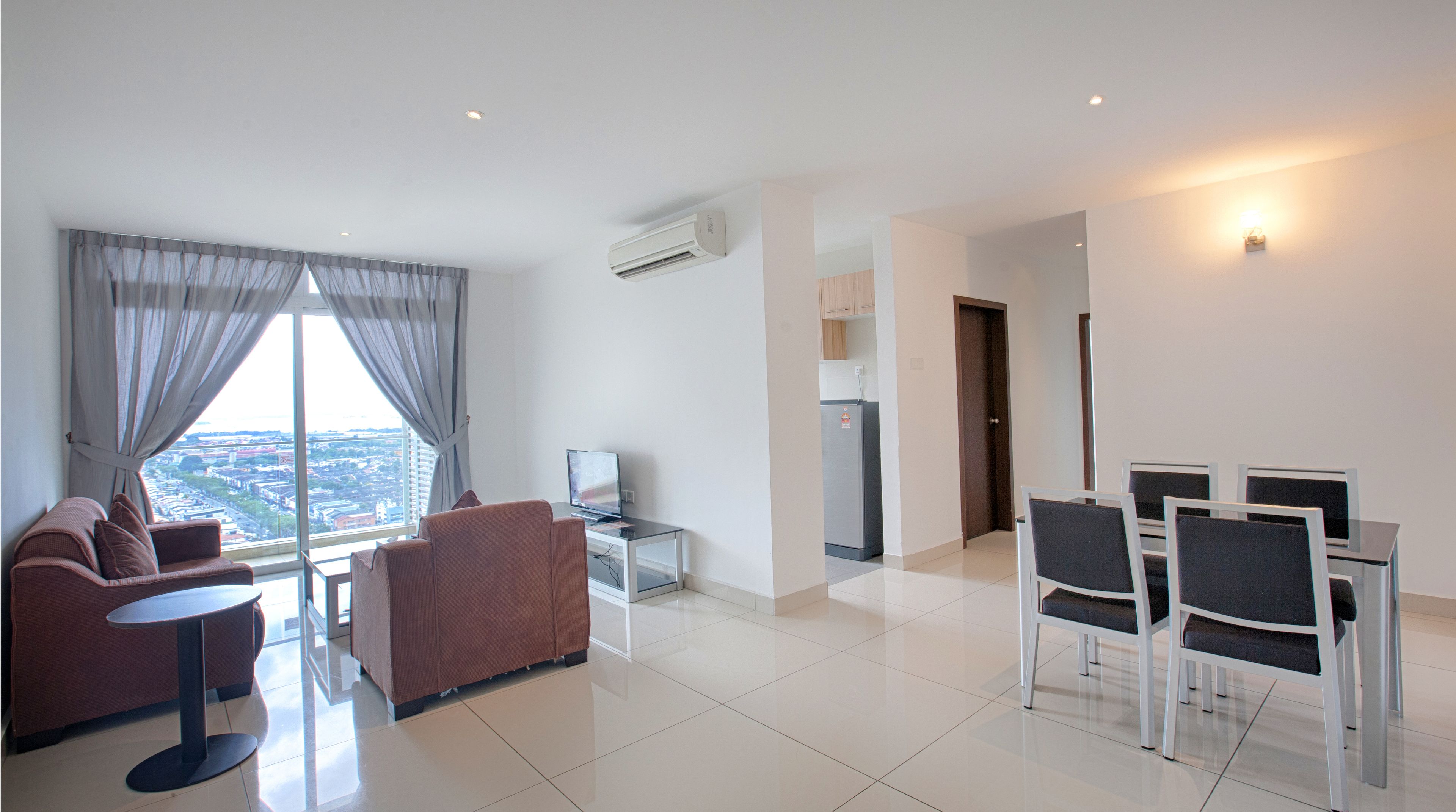 A modern living room with a sofa, a small table, and a TV. There is a kitchen area with a refrigerator and a dining area with chairs.