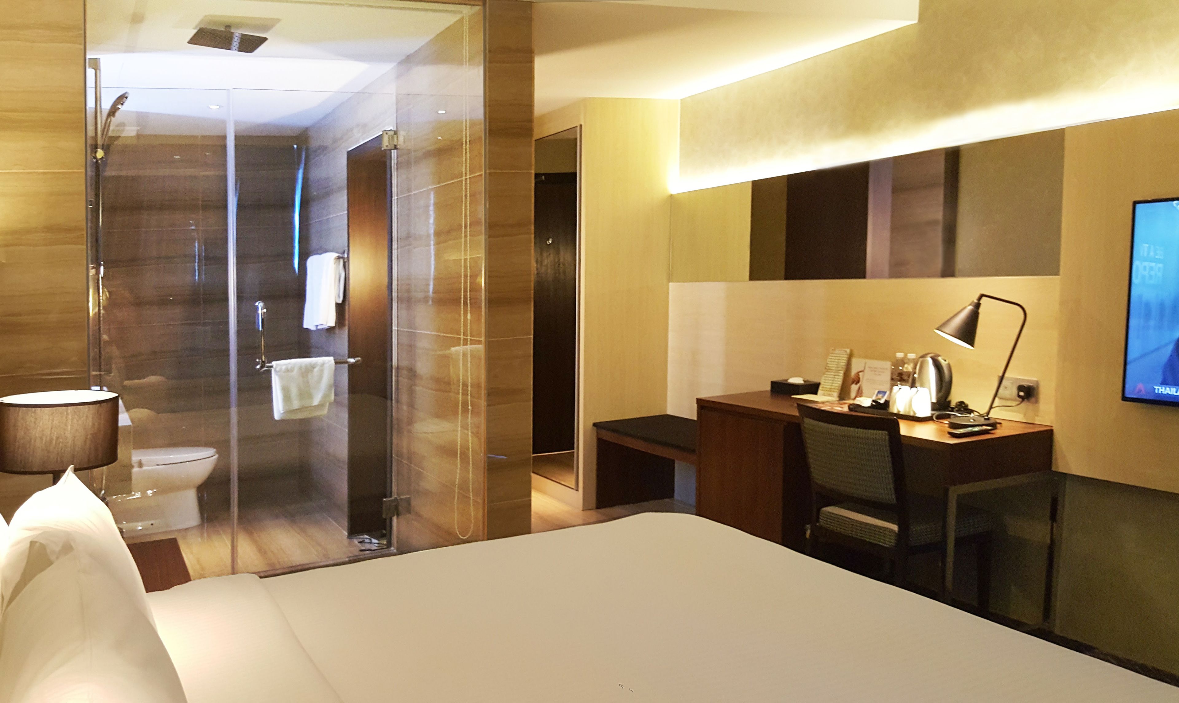 A hotel room with a bed, a desk with a chair, a desk lamp, a television, and a shower area.